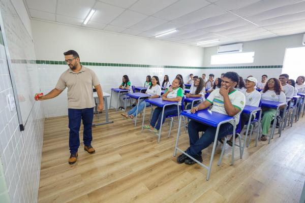 Um professor escreve em um quadro branco enquanto os alunos sentam-se em carteiras, envolvidos e sorridentes em uma sala de aula iluminada.(Imagem:Divulgação)